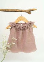 The Amayo Blossom Ruffle Dress, featuring a mauve A-line silhouette and ruffled collar, hangs on a wooden hanger from a branch. White flowers adorn the lower left corner, set against a plain light background.