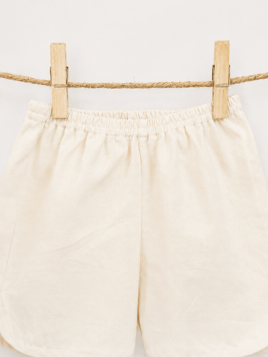 Amayo’s Oak Shorts in a cream color hang on a rope with two wooden clothespins, set against a plain white background.