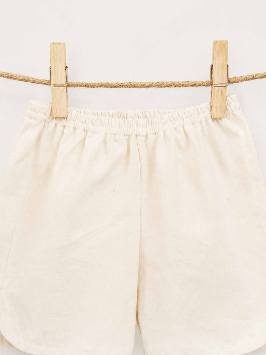 Amayo’s Oak Shorts in a cream color hang on a rope with two wooden clothespins, set against a plain white background.