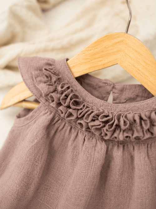 A close-up of the Blossom Ruffle Dress by Amayo, featuring a mauve A-line silhouette with a ruffled neckline, displayed on a wooden hanger. A softly blurred cream fabric appears in the background.