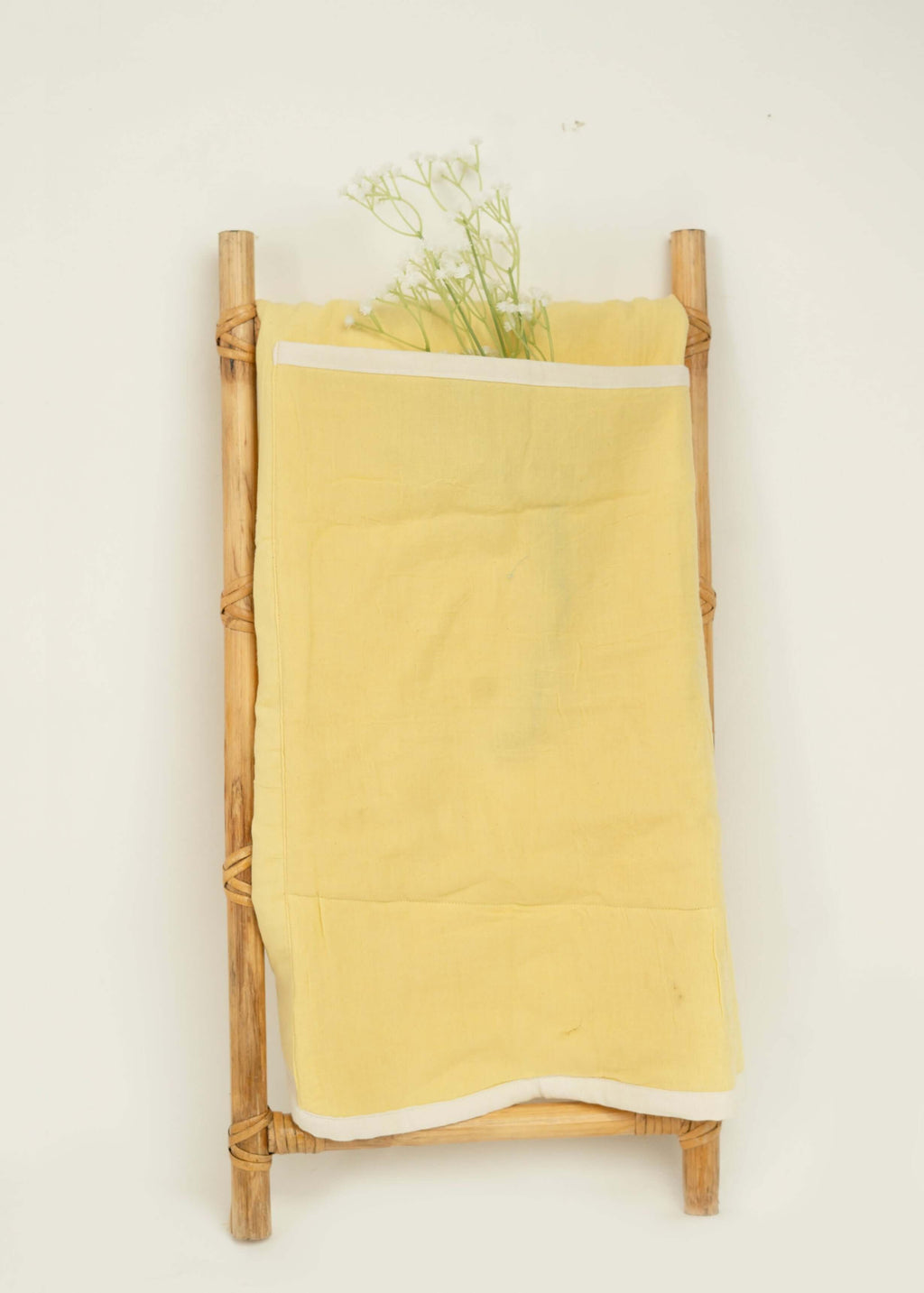 A small wooden ladder leans against a white wall, draped with the Amayo Halo Baby Holder in soft yellow organic cotton. Delicate white flowers are tucked into the fabric, creating a simple, minimalist display that evokes a cocoon of comfort.