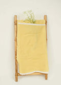 A small wooden ladder leans against a white wall, draped with the Amayo Halo Baby Holder in soft yellow organic cotton. Delicate white flowers are tucked into the fabric, creating a simple, minimalist display that evokes a cocoon of comfort.