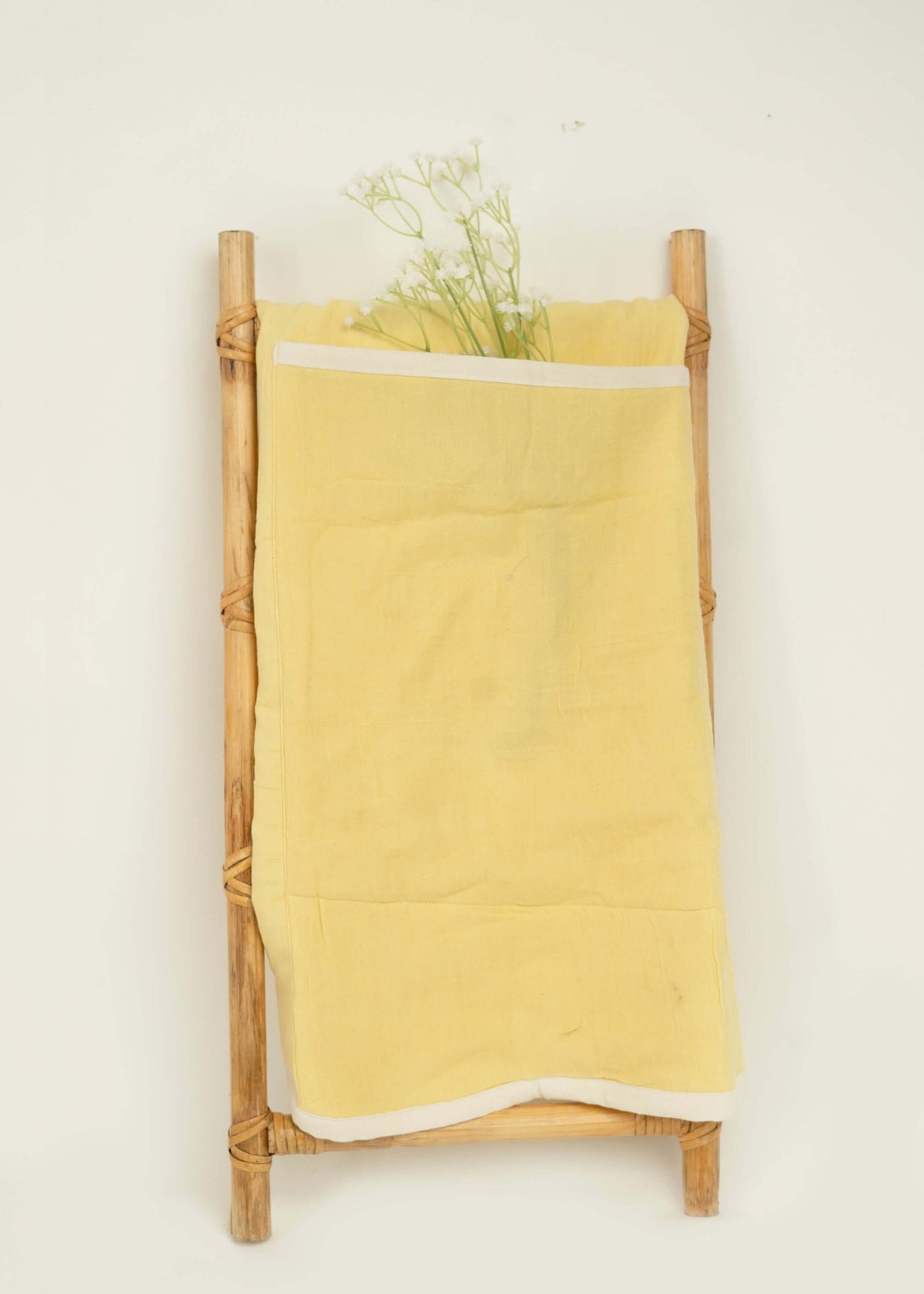 A small wooden ladder leans against a white wall, draped with the Amayo Halo Baby Holder in soft yellow organic cotton. Delicate white flowers are tucked into the fabric, creating a simple, minimalist display that evokes a cocoon of comfort.