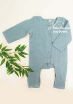 Light blue Amayo Snug Romper with long sleeves and nickel-free steel snap buttons. This cozy baby romper, designed for comfort and breathability, is displayed flat beside a sprig of green leaves on a cream background. Text highlights the easy-to-use snap buttons—ideal for your little one’s everyday wear.