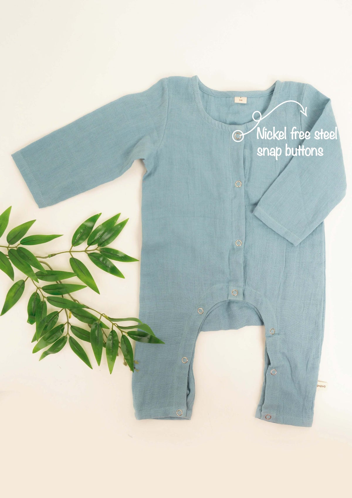 Light blue Amayo Snug Romper with long sleeves and nickel-free steel snap buttons. This cozy baby romper, designed for comfort and breathability, is displayed flat beside a sprig of green leaves on a cream background. Text highlights the easy-to-use snap buttons—ideal for your little one’s everyday wear.