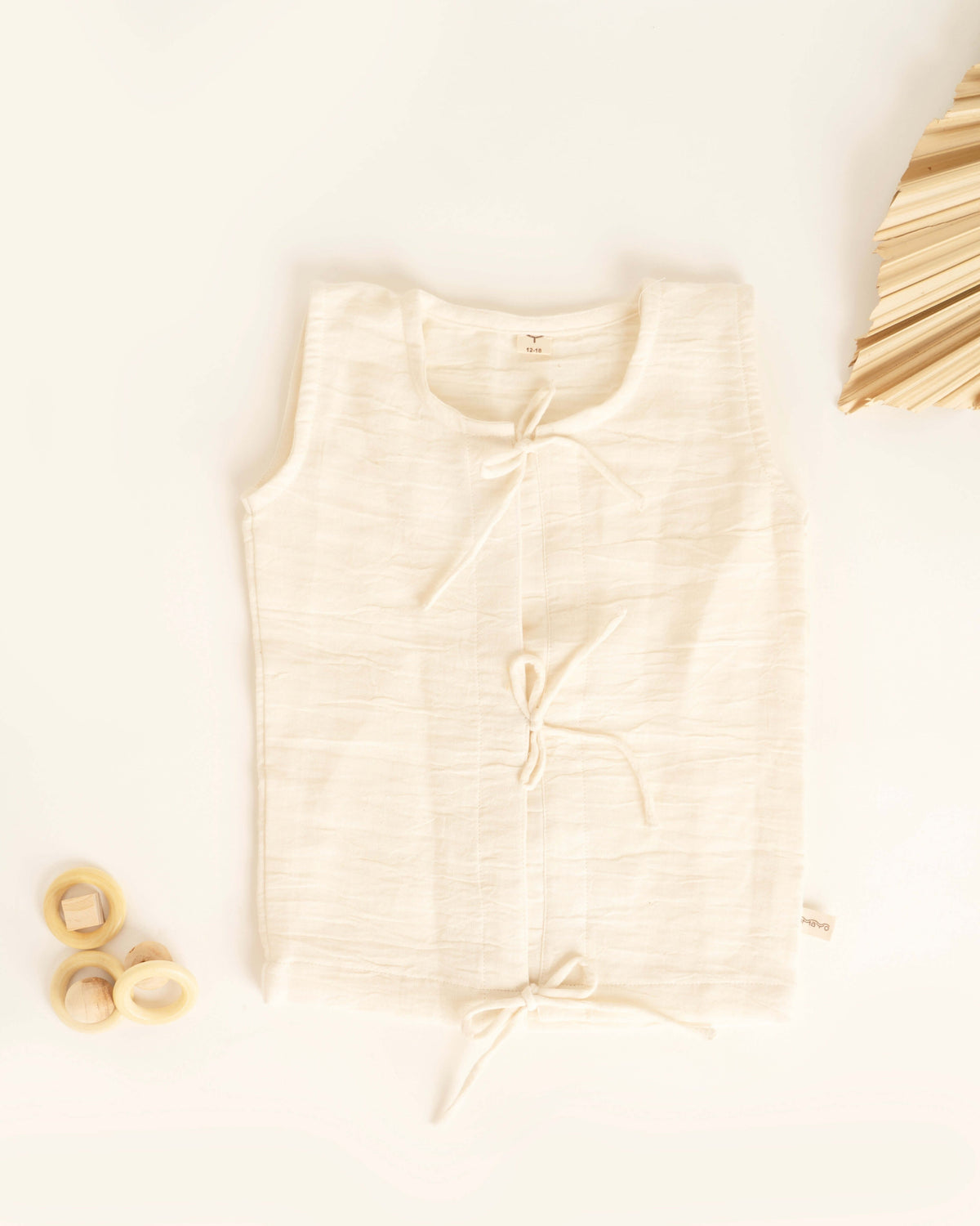 The Little Bud Vest by Amayo is a sleeveless, cream-colored baby vest crafted from organic muslin with a textured finish. This breathable piece features three front tie closures and is displayed flat on a white surface, accompanied by wooden rings and a folded beige fan for decoration.
