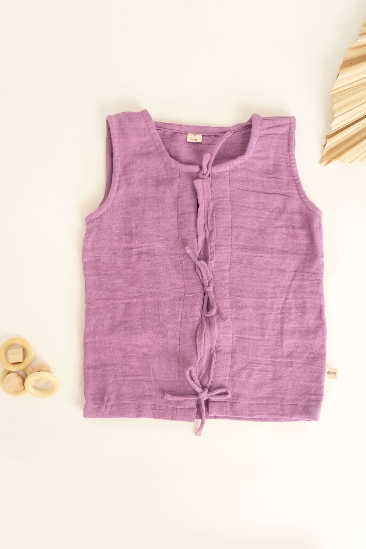 An Amayo Little Bud Vest, crafted from organic muslin in a sleeveless, light purple design with three front tie closures, is displayed flat on a white surface. Decorative wooden rings and dried palm leaves accentuate the vest’s breathable fabric and natural appeal.