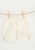The Amayo Oak Shorts, cream-colored and comfortable with an elastic waistband, hang from a rope clothesline secured by two wooden clothespins against a plain white background.