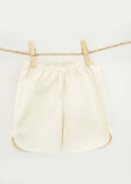The Amayo Oak Shorts, cream-colored and comfortable with an elastic waistband, hang from a rope clothesline secured by two wooden clothespins against a plain white background.