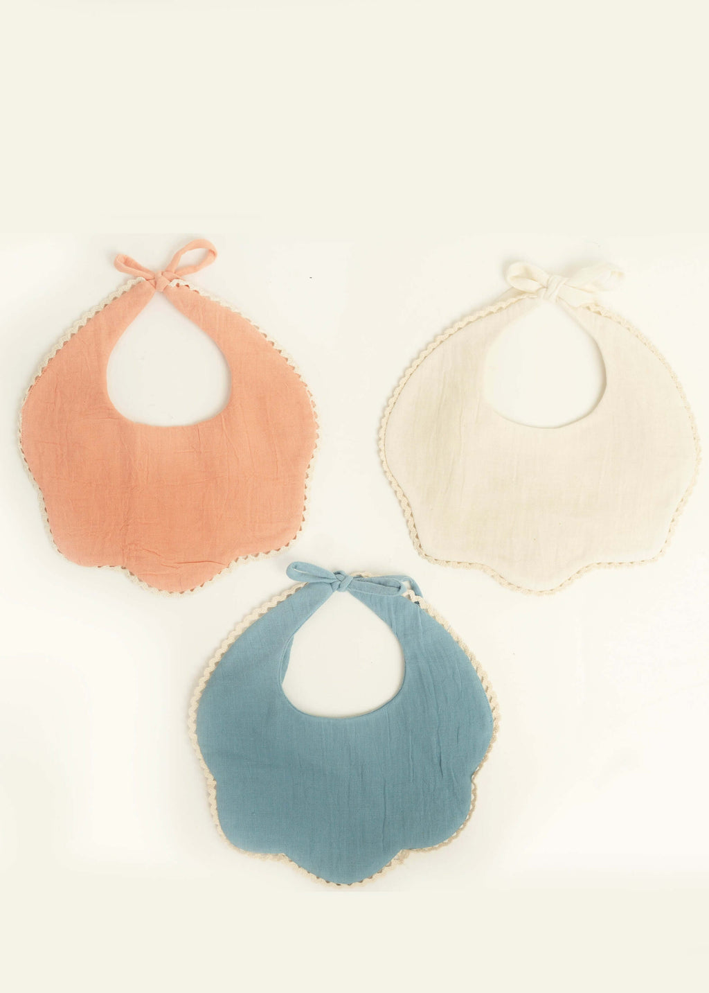 Three scalloped-edge bibs from the Amayo Petal Bib Set (set of 3) in peach, cream, and blue are arranged in a triangle shape on a light background. Each organic cotton bib features a washable design with neck ties and decorative trim along the edges, making this Amayo baby bib set both practical and charming.