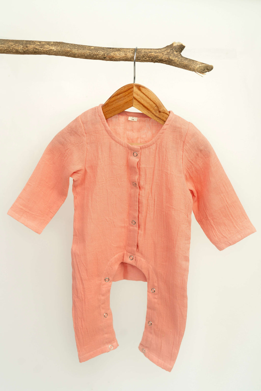 A long-sleeve, light pink Snug Romper by Amayo hangs on a wooden hanger from a branch against a plain white background. Designed from breathable baby clothing materials, this cozy romper features buttons down the front and legs to keep your little one comfortable all day.