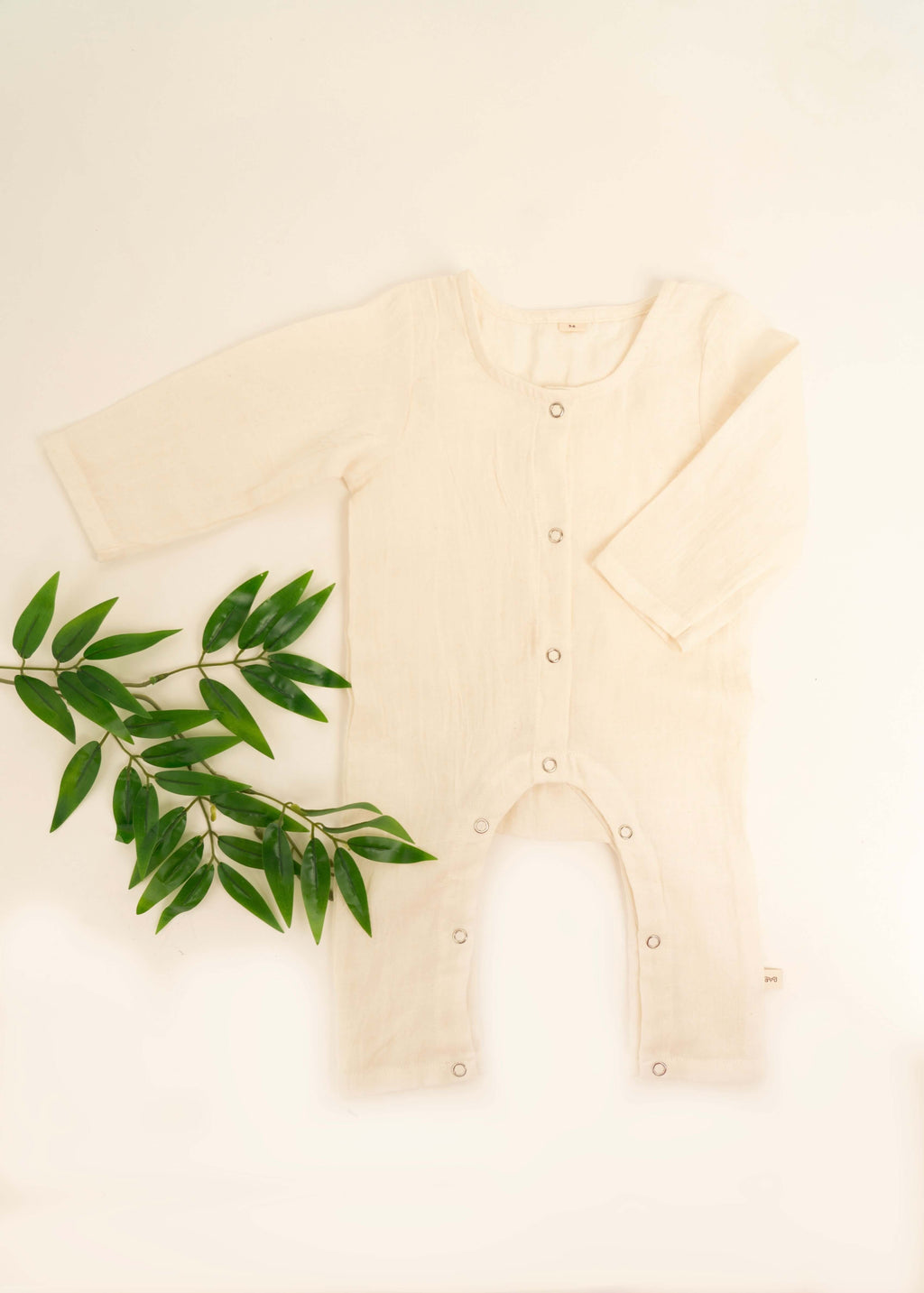 The Amayo Snug Romper, shown in a cozy cream color with long sleeves and front buttons, is laid flat on a white background beside a sprig of green leaves. Crafted for comfort and breathability, this baby clothing keeps your little one snug all day long.