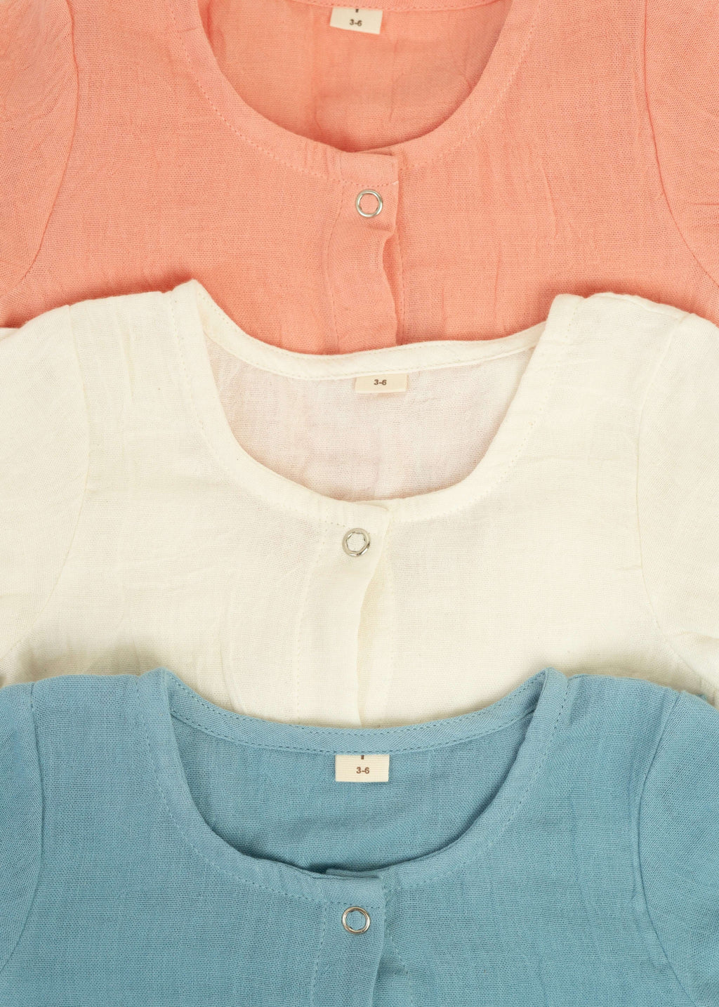 Three Snug Rompers by Amayo in pastel shades—peach, white, and blue—are neatly stacked. Each romper features a round neckline, a small button, and a size tag marked 3-6. Crafted from soft, textured fabric, these rompers are perfect examples of breathable baby clothing that keep little ones comfortable throughout the day.