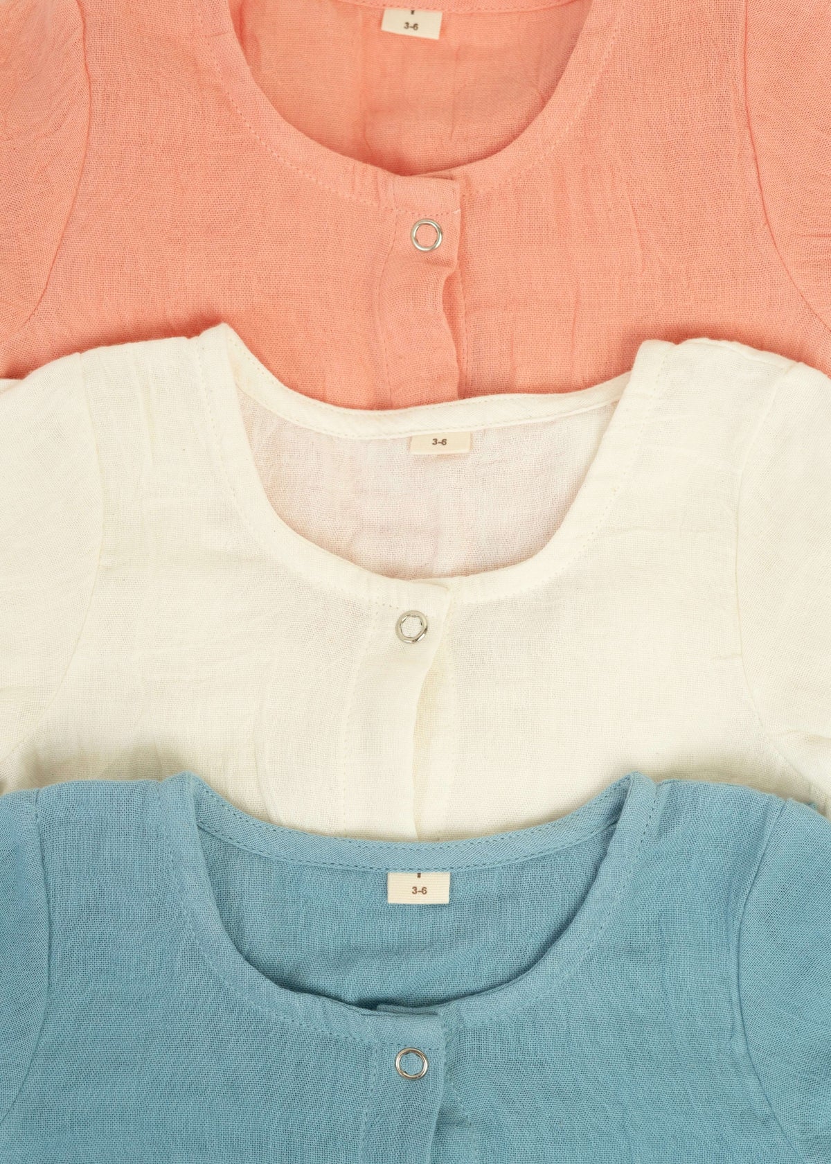 Three Snug Rompers by Amayo in pastel shades—peach, white, and blue—are neatly stacked. Each romper features a round neckline, a small button, and a size tag marked 3-6. Crafted from soft, textured fabric, these rompers are perfect examples of breathable baby clothing that keep little ones comfortable throughout the day.