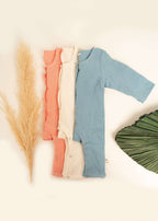 Three long-sleeve Snug Rompers by Amayo in coral, cream, and blue are neatly arranged next to dried pampas grass and a large green leaf on a cream background, offering breathable baby clothing for your little one.
