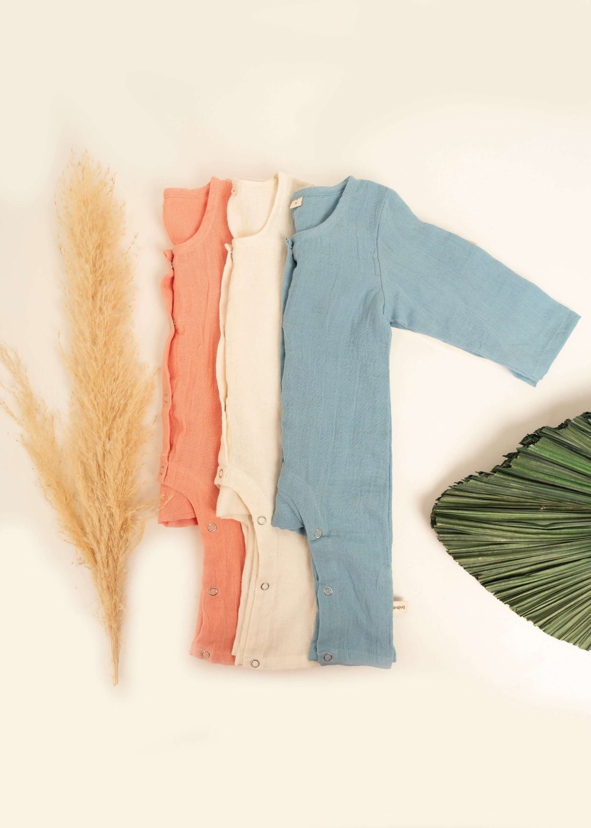 Three long-sleeve Snug Rompers by Amayo in coral, cream, and blue are neatly arranged next to dried pampas grass and a large green leaf on a cream background, offering breathable baby clothing for your little one.
